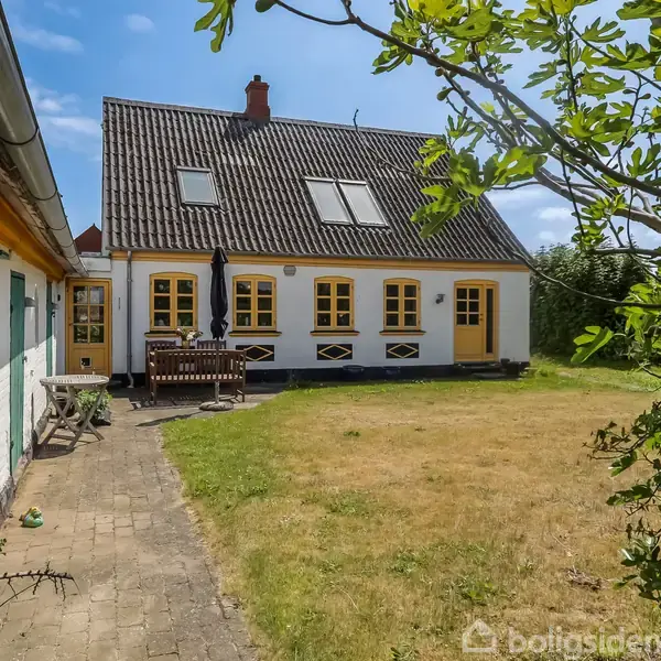 Et charmerende hvidt hus med gule skodder står i en grøn have. Der er en terrasse med havemøbler foran huset. Himmel er blå med skyfri udsyn.
