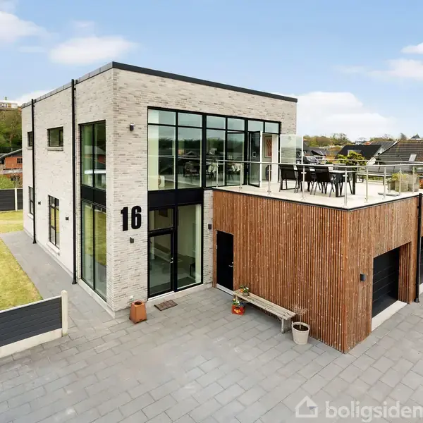 Et moderne toetagers hus med store vinduer og tagterrasse står i en boligkvarter. Husnummer "16" vises på bygningen. Omkring det er en have med græsplæne og flisebelagt gårdsplads.