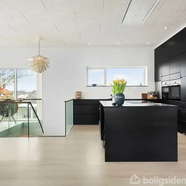 Sort køkkenø med blomster på og indbyggede skabe i et lyst rum. Store vinduer giver udsigt til et træ. Moderne design med lyse trægulve og stilrent interiør.