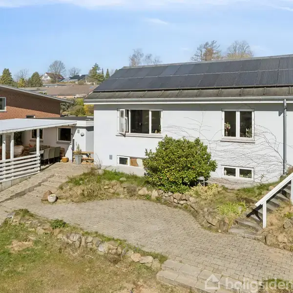 Hvidt hus med solpaneler på taget står i en have med stenbelægning og en grussti. Til venstre er en carport med tag.