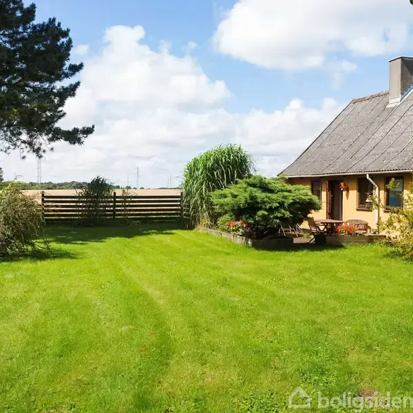Hus med stråtag står stille ved siden af en grøn have fyldt med buske og træer, omkranset af et lavt stakit og marker under en skyet himmel.