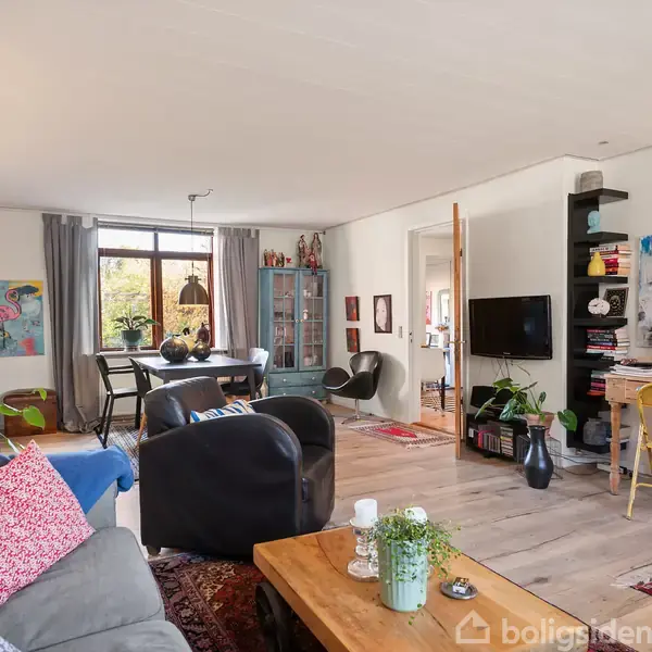 En stue med sofaer og stole står arrangeret omkring et sofabord, mens et fjernsyn er monteret på væggen. Rummet er lyst og indrettet med diverse malerier og planter.