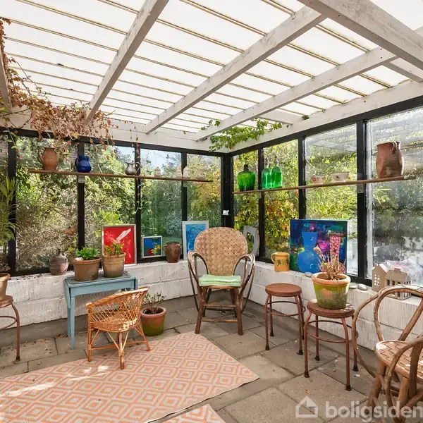 Værelse med glasvægge fyldt med stole og borde, dekoreret med planter og farverige malerier, placeret på opbevaringshylder. Grønne træer kan ses udenfor gennem vinduerne.