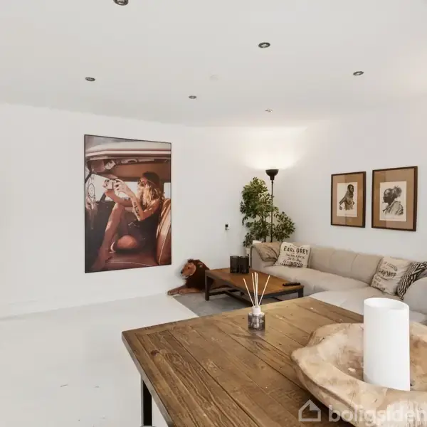 Et moderne stue med hvide vægge, hvor en stor sofa er placeret i hjørnet. Et rundt træbord med en hvid lysestage og dekorationer står i forgrunden. Væggen er dekoreret med billeder og en plakat af en kvinde i en bil. Grønne planter tilføjer farve og atmosfære.