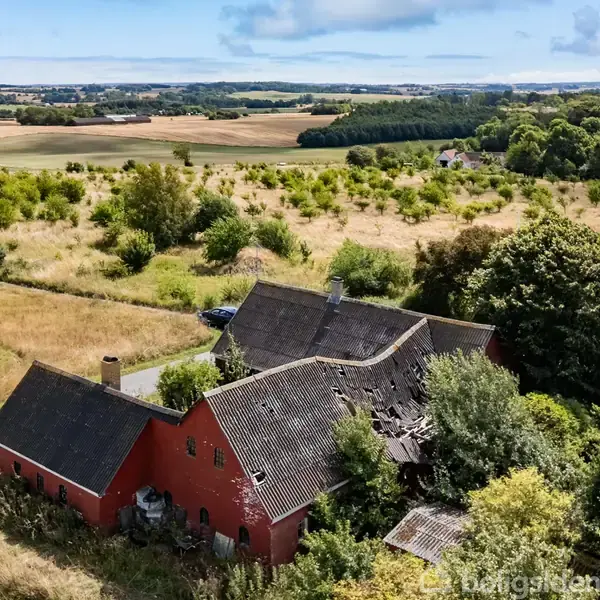 En rød murstensbygning med skråt tag står delvis forfaldent i et åbent landligt landskab. Omgivelserne består af marker, træer og en landevej.