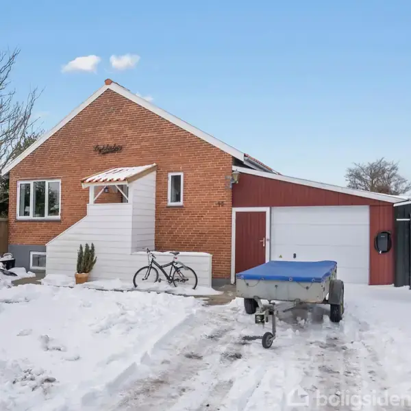 Et rødt murstenshus med hvidt indgangsparti har en cykel og trailer foran; omgivende sne og en blå himmel skaber en vinterstemning.
