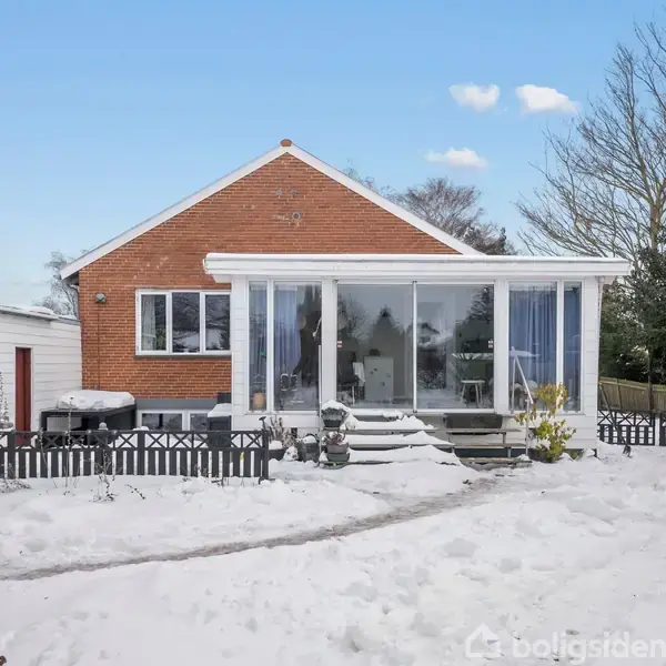 Et rødt murstenshus med store vinduer ligger i en snedækket have. Sne dækker også tag og omgivelser, mens træer uden blade omkranser ejendommen i baggrunden.