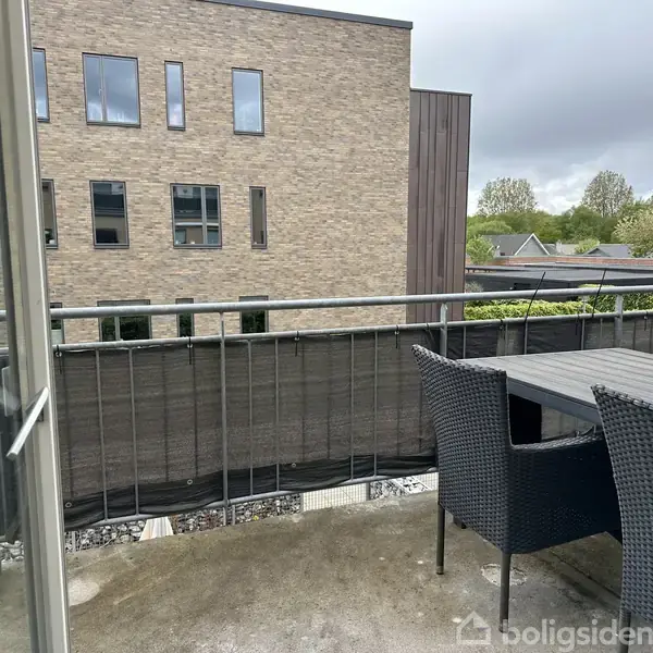 Et udendørs balkonområde med kurvemøbler står stille. I baggrunden ses en moderne murstensbygning omgivet af grønne trætoppe og en grå himmel.