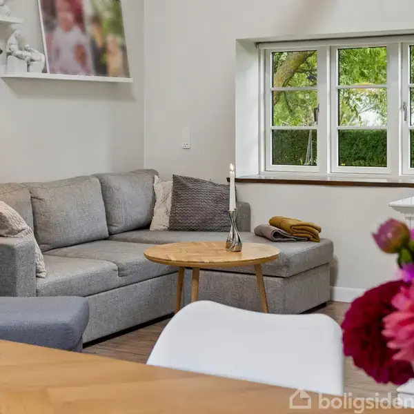 Grå sofa med pyntepuder står ved siden af et vindue med udsigt til grønne træer. Rundt bord med lysestage. Blomster i forgrunden på et træbord.