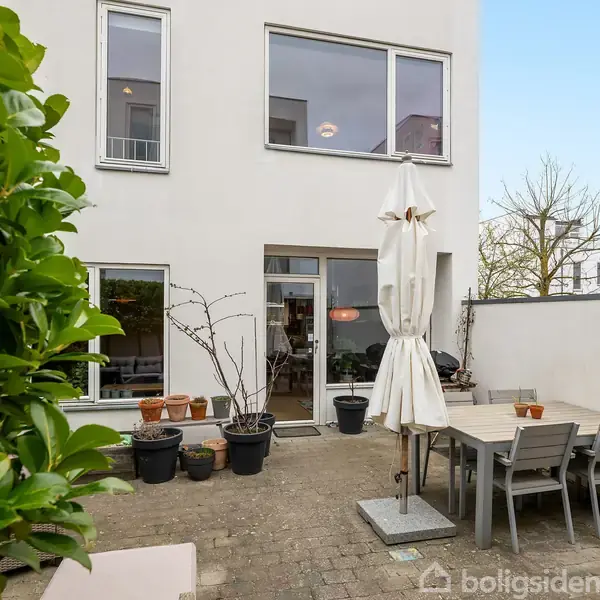 En udendørs terrasse har havemøbler og en parasol. Omgivet af en hvid bygning med store vinduer og blomsterpotter, ligger området i en urban havekontekst.
