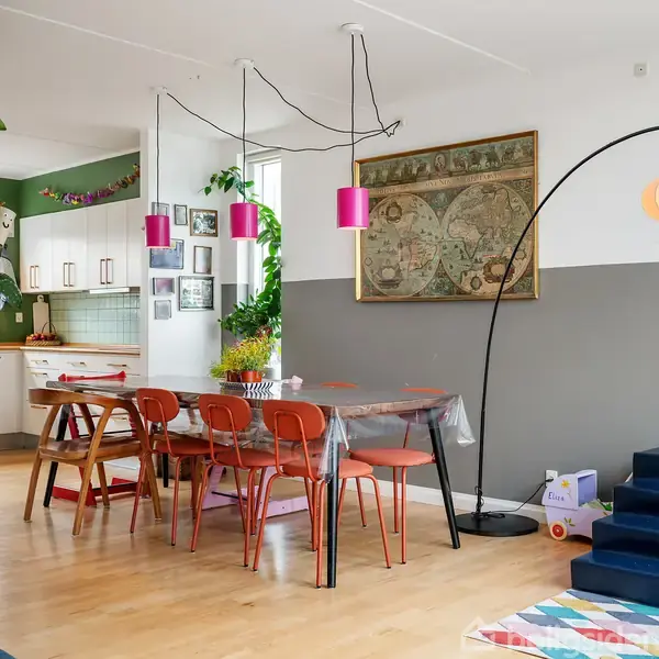 Spisebord med orange stole er omgivet af farverige lamper og en stor gulvlampe i en indretningsvenlig spisestue. I baggrunden ses et åbent køkken med hvide skabe.