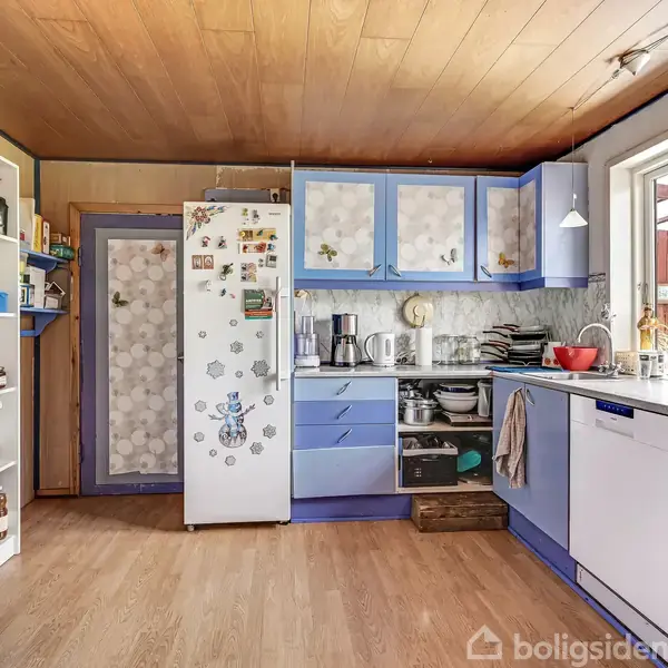 Køleskab med magneter står i et blå-hvidt køkken, omgivet af skabe, hylder og en opvaskemaskine under en stor vindue, hvor der også er placeret tallerkener og køkkenredskaber.