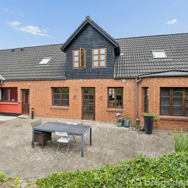 Et rødt murstenshus med sorte tagsten har en gårdsplads foran. Der står et bord og to stole på brostenene. Huset har flere vinduer og en kvist med træbeklædning.