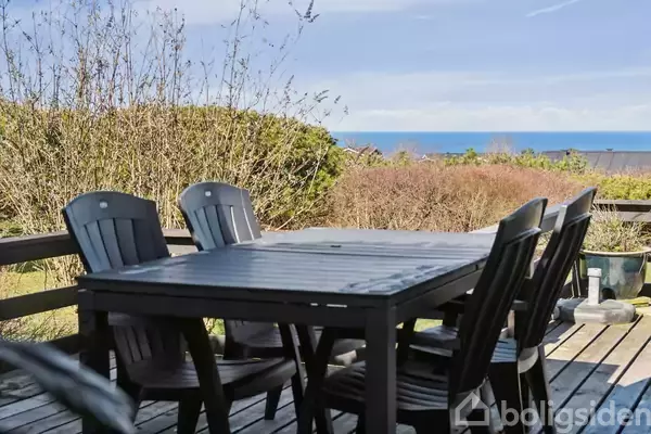 En udsigt viser tætte buske og et tag i forgrunden, med havet og en blå himmel i baggrunden, under en klar dag.