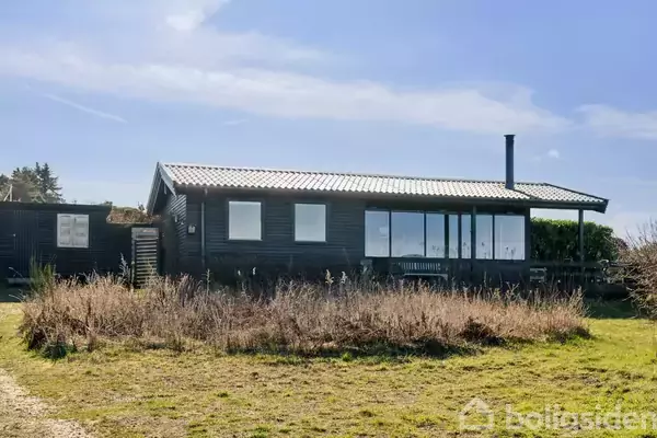 Et sort havebord med seks stole står stille på en træterrasse. Omgivet af buske og udsigt til havet i horisonten under en blå himmel.