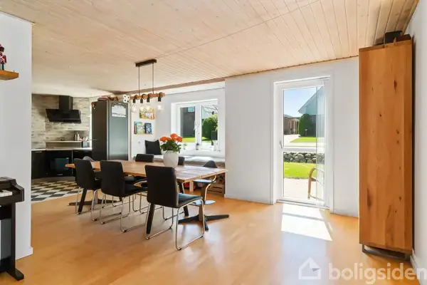 Trappe med sort gelænder og trætrin snor sig opad. Entré med trægulv, hvidmalede vægge og træbeklædt loft. Skohylde og knagerække ses på væggen. En dør fører videre ind.