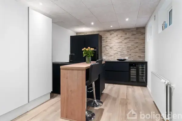 En sort sofa står i en lys stue med trægulv, ved siden af et stort vinduesparti. Et træbord med blomster i midten, omgivet af vægreoler og udsigt til en have.