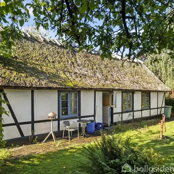 Bindingsværkshus med stråtag står i en grøn have. Flere vinduer ses langs muren, og et fuglehus og havemøbler er placeret foran huset. Sollys skinner gennem grenene.
