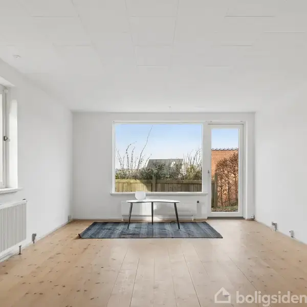 Et enkelt bord står på et tæppe i et lyst rum med trægulv. Store vinduer giver udsigt til en have. Vægge og loft er hvide.