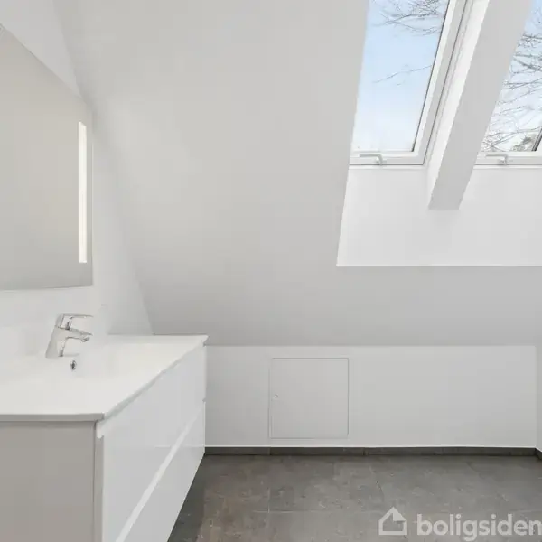 Hvidt badeværelse med skrå loft og to ovenlysvinduer. Vask med spejl står mod venstre væg. Flisebelagt gulv og minimalistisk design skaber et lyst og rummeligt indtryk.