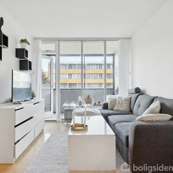 Grå sofa står i en moderne stue med hvide vægge, store vinduer og et tv på et lavt skab. Der er hylder på væggen og en glasdør til en balkon.