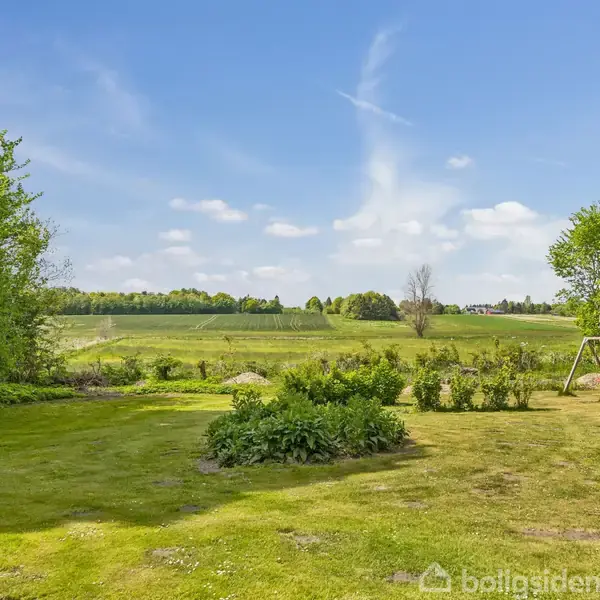Græsplæne med gyngestativ til højre i et åbent landskab omgivet af træer og buske. Skyhimmel med lette skyer i baggrunden.
