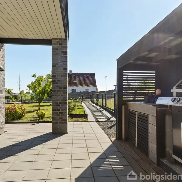 Grill i en sort pergola, står stille ved siden af en moderne terrasse med flisebelagt gulv, omgivet af en velplejet have med blomster og grønne træer i baggrunden.