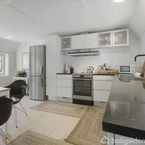 Køkken med ovn og køleskab, moderne indretning. Bord med stole ved vindue, lys træ- og sort farveskema. Lys strømmer ind fra vinduer. Tæpper på gulvet.