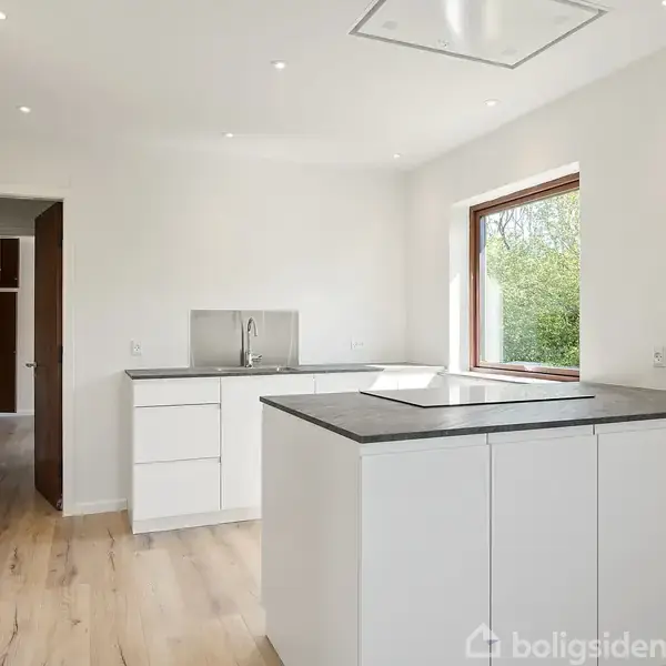 Køkken med hvide skabe og en grå bordplade står i tomt, lyst rum. En stor vindue til højre lader naturligt lys ind med udsigt til grønne områder.