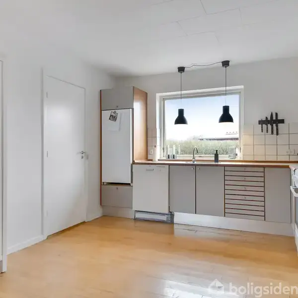 Køkkenet har et lysindfald fra vinduet og indeholder et hvidt skab med service, et køleskab, og vask. To sorte lamper hænger over køkkenbordet med træoverflade.