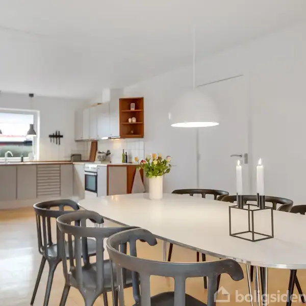 Et spisebord med sorte stole står i et rummeligt køkken-alrum. På bordet er en vase med blomster og en lysestage. Rummet er lyst med træloft og moderne indretning.