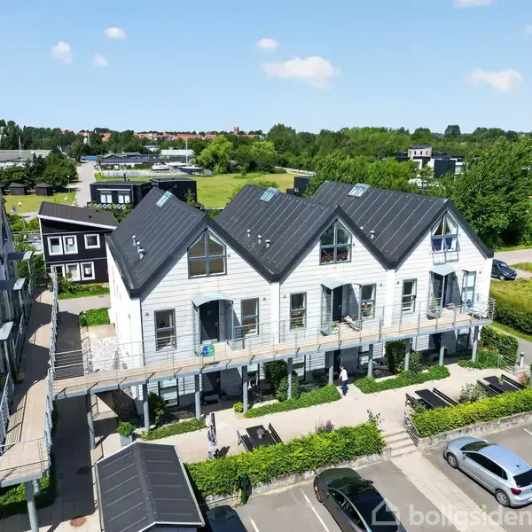 Flerradet, moderne bygning med skrå tage og store vinduer. Bygningen ligger i et grønt område med andre lignende bygninger i nærheden. personer ses foran bygningen.