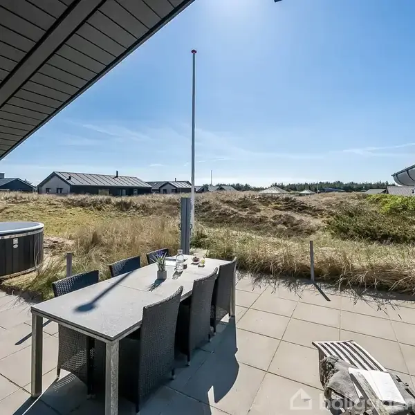 En udendørs terrasse med et spisebord og seks stole står under et halvt overdække. En flagstang og en hot tub er i nærheden. Omkringliggende klitter kan ses under en klar blå himmel.