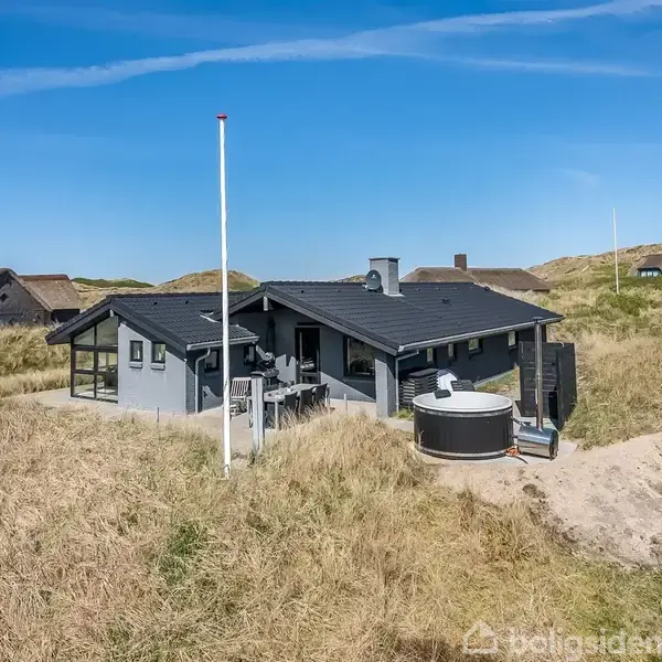 Sommerhus stående på sandklitter, med et flagstang i nærheden. En udendørs jacuzzi er synlig ved siden af huset. Flere klitter og huse i baggrunden under en blå himmel.