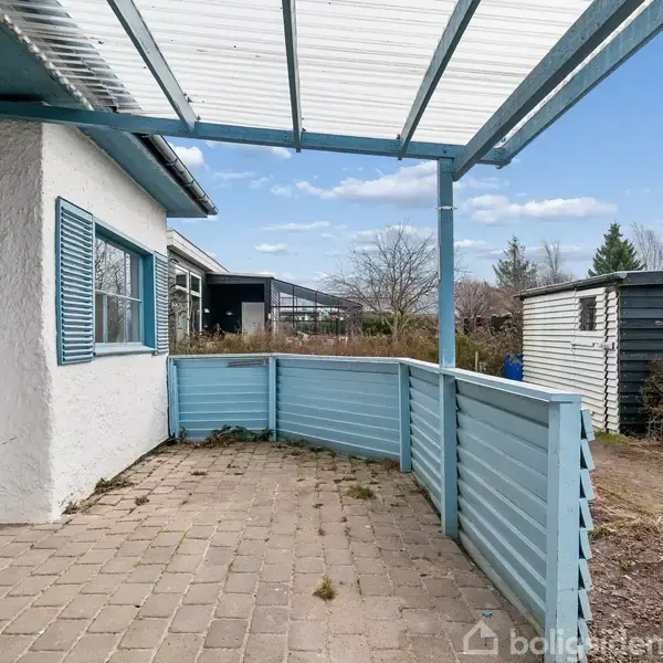 Terrasse med blåmalet rækværk og overhæng, tilstødende hvidt hus med lyseblå skodder. Omgivet af have og træer, med skur i baggrunden. Klar blå himmel over området.