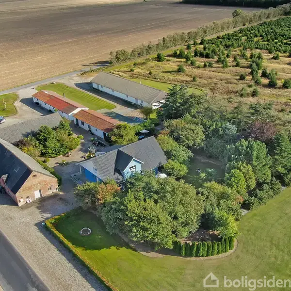 En gård med adskillige bygninger omkranset af frodige træer og store marker. Der er en vej langs gården, og området er præget af landlig idyl og åbne landskaber.