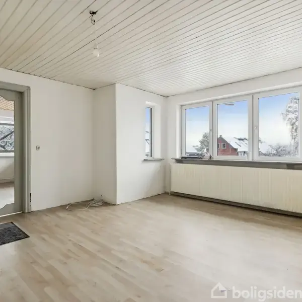 Et tomt rum med trægulv og hvide vægge. Store vinduer lader naturligt lys komme ind, og en glasdør fører til en anden del af boligen. En radiator ses under vinduerne.