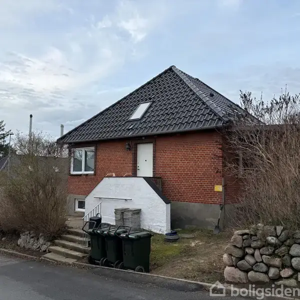 Rødt murstenshus med sort tag står i villakvarter. Enkeltstående dør øverst på trappe, omgivet af vinduer. Fortov foran med affaldscontainere ved trappe. Lav stenmur og buske rundt om.