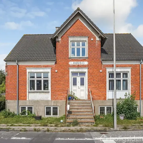 Et rødt murstenshus med gråt tag har inskriptionen "HØJBO" over hoveddøren. Huset står bag trapper, omgivet af en lille have og en vejbane foran.