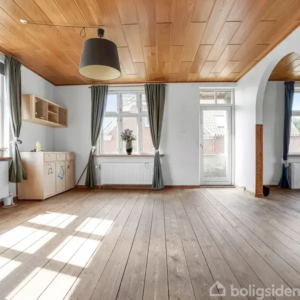 Træbeklædt rum med brune trægulve og store vinduer, der slipper lys ind. En stor lampe hænger fra loftet. Gardiner omgiver vinduerne og en dør fører ud.