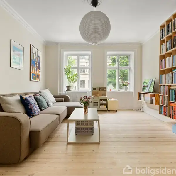 Stue med en stor grå sofa til venstre, dekoreret med puder. Et sofabord med blomster står centralt. Højre væg har en stor bogreol. To vinduer giver naturligt lys.