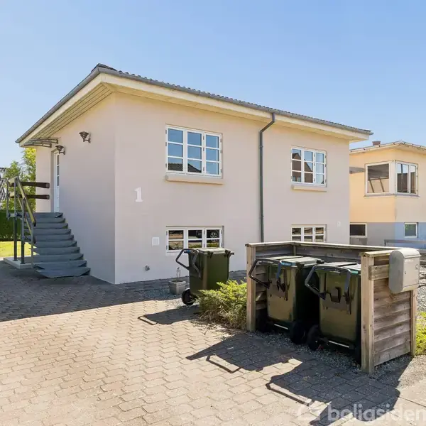 Et lyst hus står i en villakvarter. Huset har store vinduer og en trappe ved siden. I forgrunden er affaldscontainere ved en flisebelagt indkørsel.
