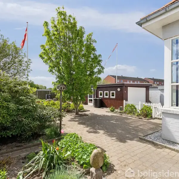 En velplejet have med blomsterbede omgivet af buske og træer, en flisebelagt sti og lille brunt skur i baggrunden. Et rødt dansk flag er synligt i haven.