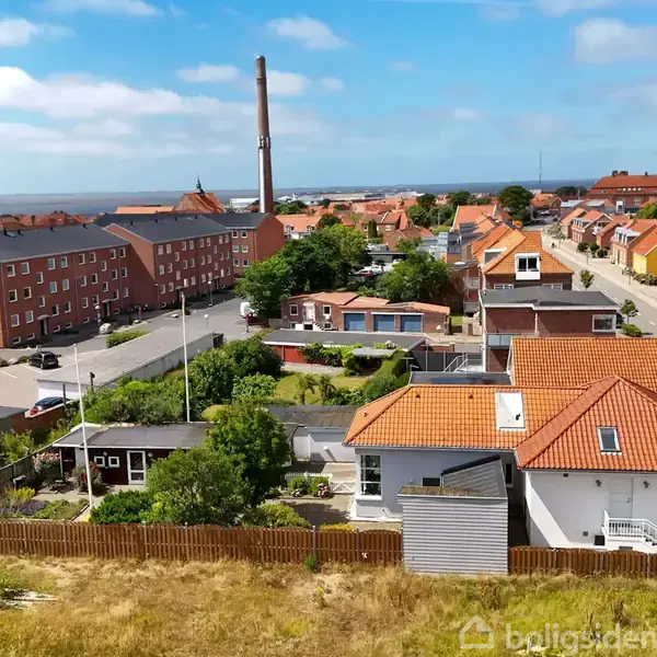 Et hus med rødt tag og hvide vægge står stille i en boligområde; omgivet af flere bygninger, græs og træer under en klar, blå himmel.