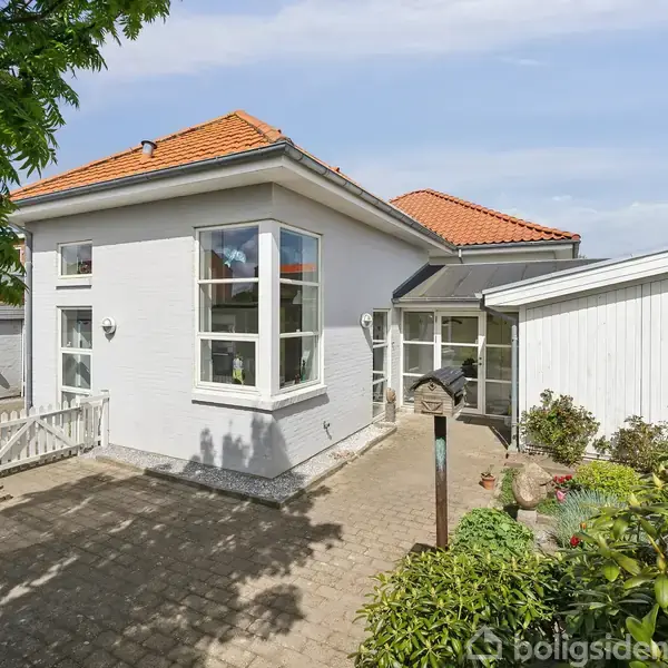 En hvidmalet villa med rødt tegltag ligger i en solrig have. Huset har store vinduer og en gårdsplads med brostensbelægning omgivet af grønne buske og nogle træer.