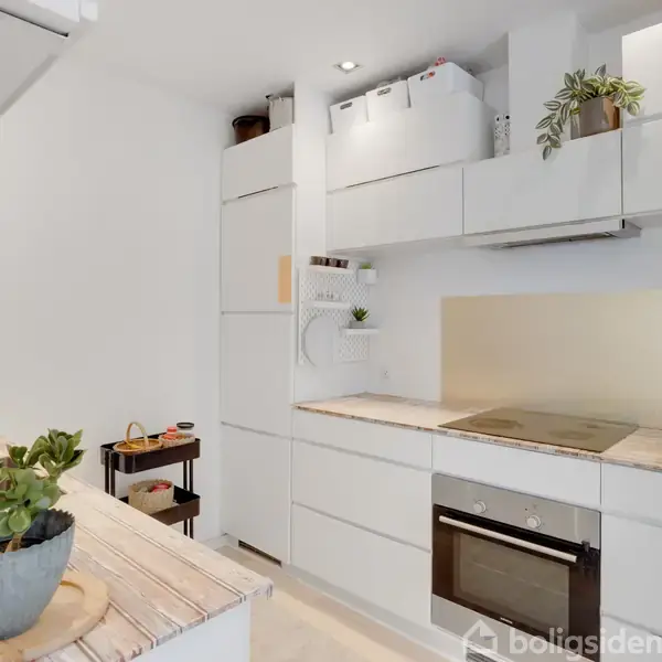 Et moderne køkken med hvide skabe, en indbygget ovn og kogeplade til venstre. En plante er placeret på bordet. Lys og stilren indretning.