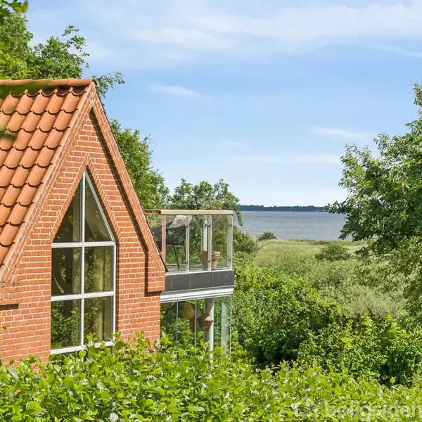 Murstenshus med rødt tegltag står i sollys omgivet af frodig grøn vegetation. I baggrunden ses en sø, hvilket skaber en naturskøn udsigt.