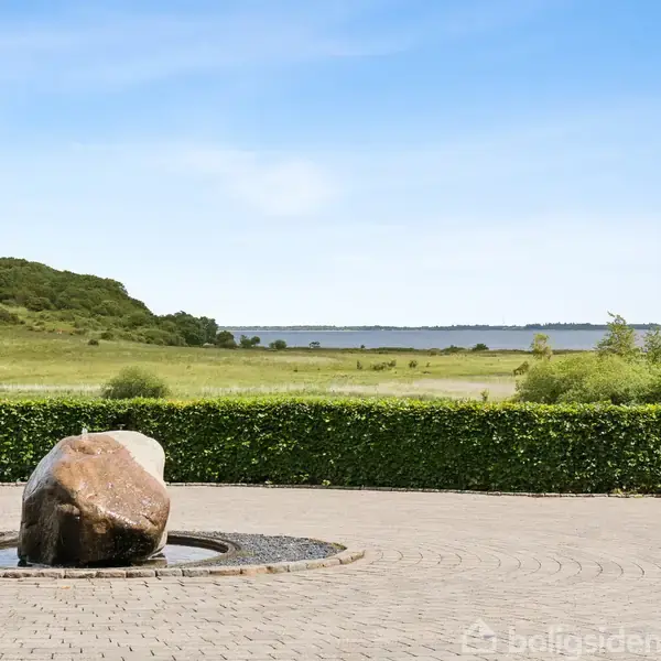 Stor sten ligger centralt i en rund vandinstallation på en brolagt plads, omgivet af en lav hæk med udsigt til en grøn mark og en fjord i baggrunden.