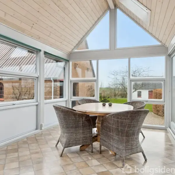 Rundt bord med flettede stole står i en lys udestue med skråt træloft og store vinduer, der giver udsigt til en have og en murstensbygning udenfor.