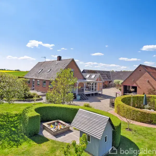 Hus omgivet af frodige grønne haver. Stier snor sig gennem området med klippede hække og træer. Klart solskin belyst landskabet, giver en indbydende og fredfyldt atmosfære.